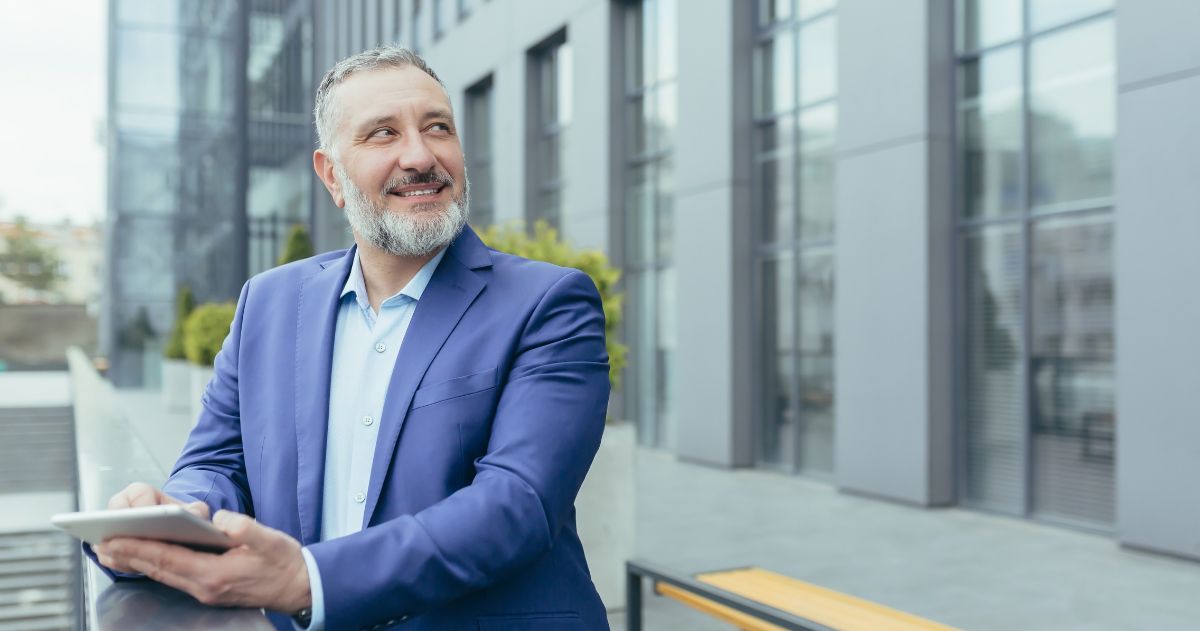 Coordenador de Programa de Pós-Graduação - Professor sênior do sexo masculino, de pé na varanda de um campus, segurando um tablet